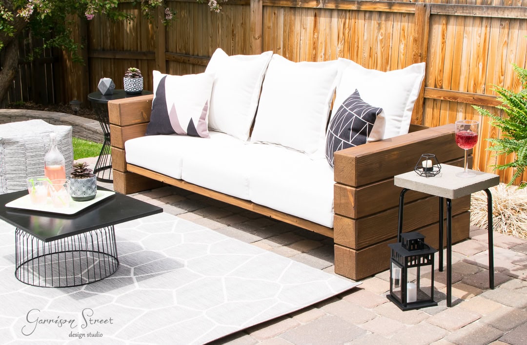 A cozy outdoor patio showcases a wooden sofa with white cushions and geometric pillows. A clever Ikea hack transforms a small table, holding a drink and lantern, while a modern coffee table with decor sits atop a patterned rug. A wooden fence and lush greenery complete the inviting scene.
