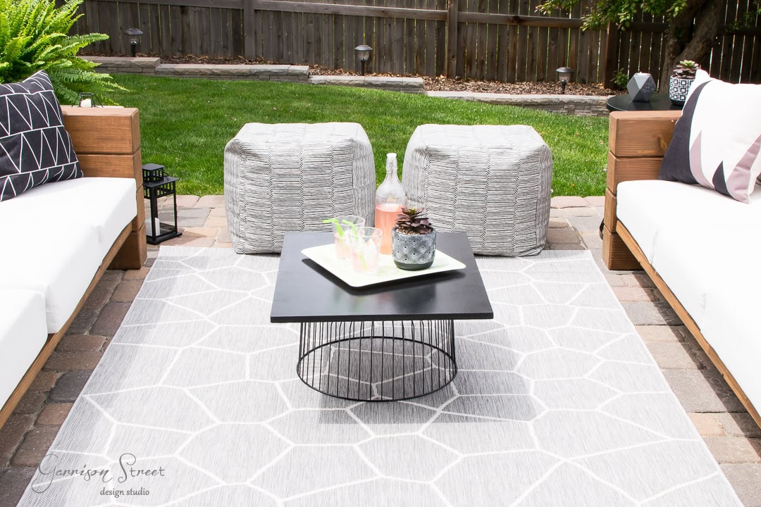 Outdoor patio with two white cushioned sofas and two gray ottomans surrounding a black square coffee table. The table holds a tray with drinks and a plant. This backyard setup is enhanced by a geometric-patterned gray outdoor rug.