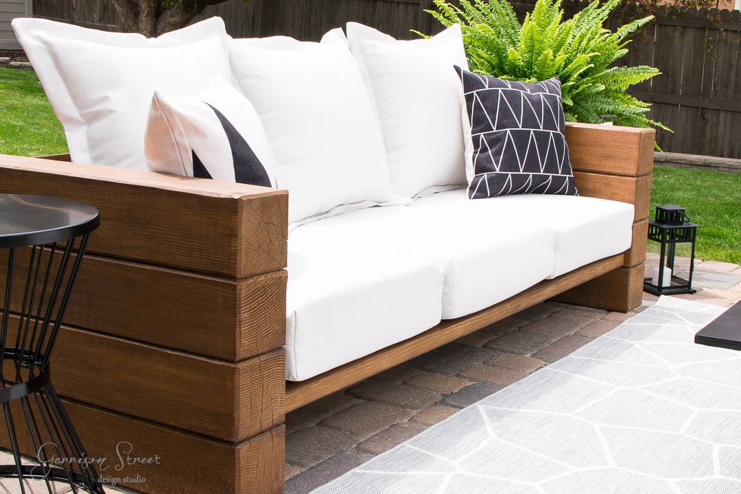 A wooden outdoor sofa with white cushions and patterned throw pillows graces the backyard patio. A small black metal side table stands beside the sofa, while lush green ferns in the background add a touch of nature.