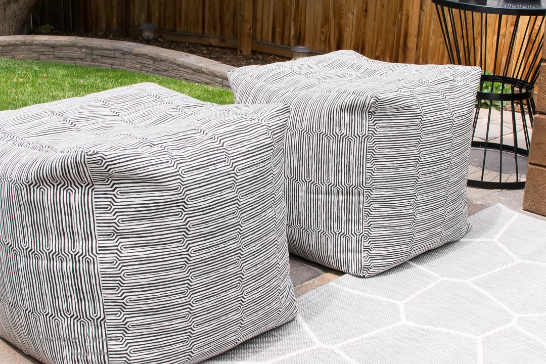 Two square poufs with a black and white geometric pattern are placed on a light gray hexagonal-patterned rug in the backyard. They are perfectly positioned on the patio area with a wooden fence and lush green grass in the background.
