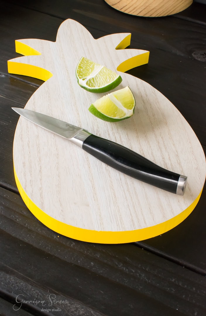 A pineapple-shaped cutting board with a yellow edge, perfect for backyard gatherings, holds a small knife and lime wedges. It rests on a dark wooden surface.