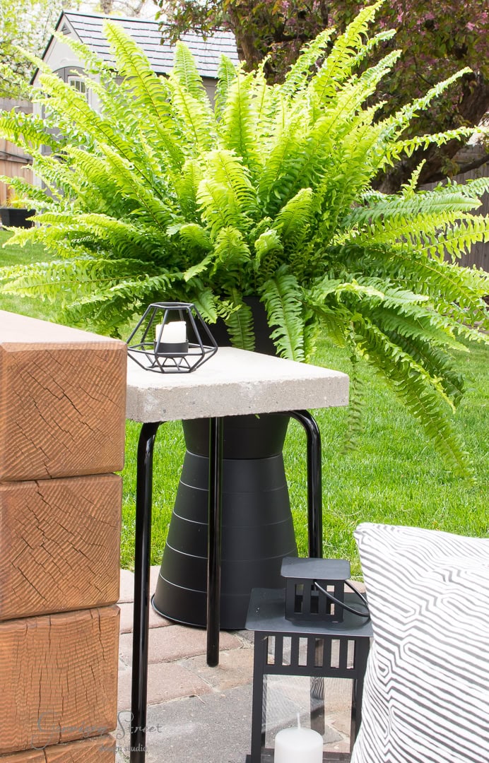 A lush fern in a black pot sits on an Ikea patio table. A geometric candle holder is beside it. Wooden beams and a striped cushion are visible, with a green lawn and trees in the background.