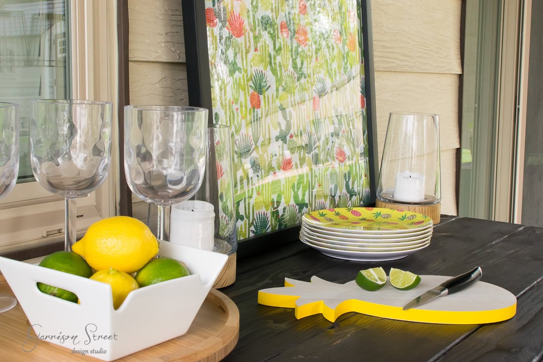 A patio table with a tropical theme features a white bowl of lemons and limes, a cutting board with lime wedges, stacked pineapple-patterned plates, and a decorative art piece with colorful tropical plants. Wine glasses and candles add elegance to the serene backyard setting.