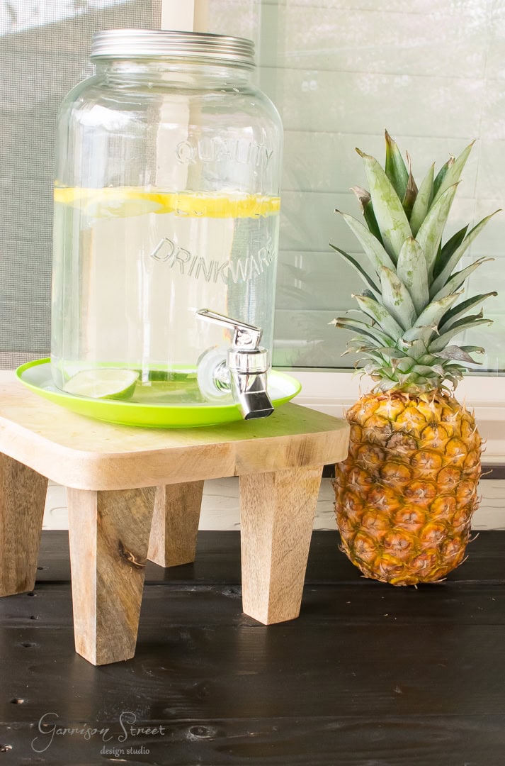 A glass drink dispenser with lemon slices sits on a wooden stand, perfect for a refreshing patio gathering. A whole pineapple adds a tropical touch to the backyard table.