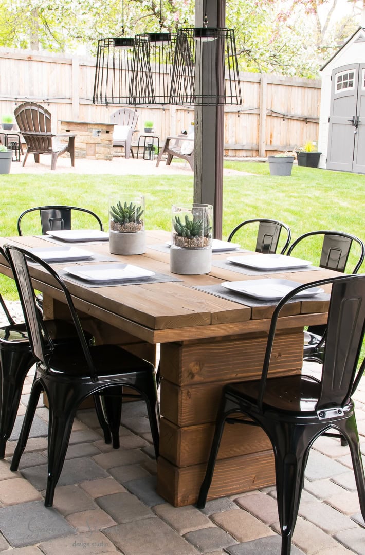 Outdoor patio dining area with a wooden table set for eight, featuring black metal chairs. The table is decorated with two succulent centerpieces. Overhead, a metal chandelier is suspended. A grassy backyard and white shed are visible in the background.