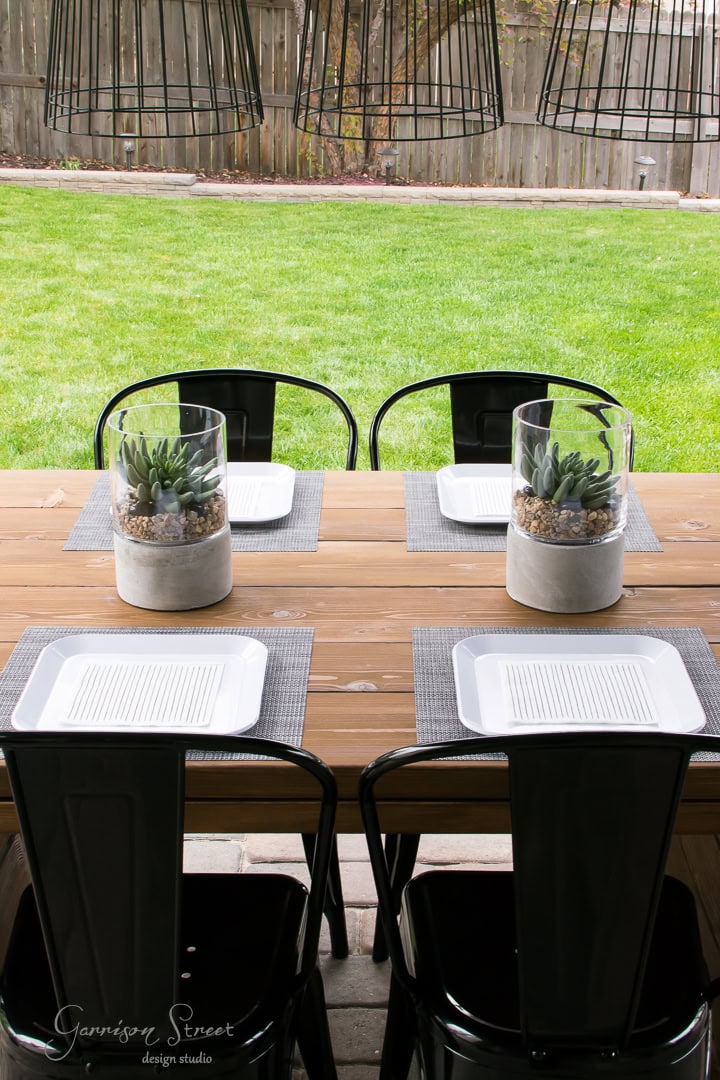 A wooden outdoor table on the patio is set for four, with black metal chairs. Each setting features a square white plate on a gray placemat. Two glass planters with succulents sit centrally, overlooking a green lawn and wooden fence in the backyard.
