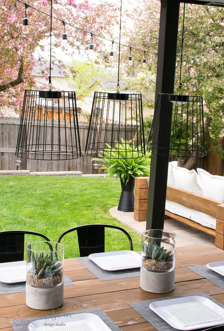 The backyard patio features a wooden table set with white plates and small succulent centerpieces. Black wire pendant lights hang above. In the background, a lush green lawn, potted fern, string lights, and blooming trees create a serene ambiance.