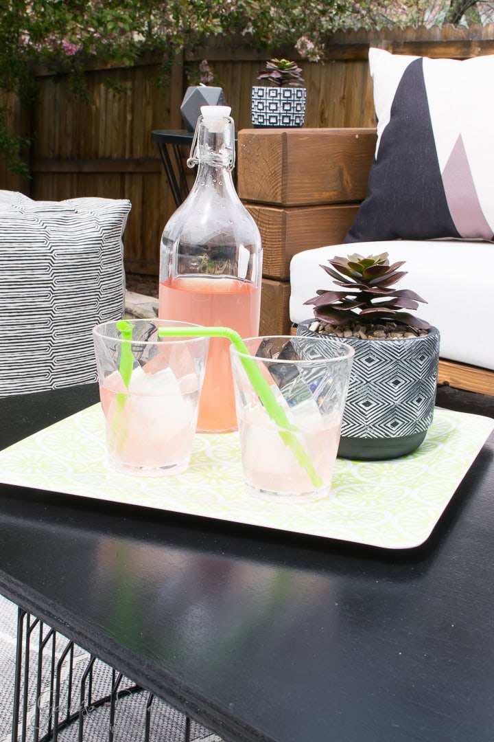 A tray with two glasses filled with a pink drink and green straws sits on a table beside a potted succulent. A bottle of the same drink is placed behind the glasses. The setting appears to be a cozy backyard patio with cushioned seats and a wooden fence.