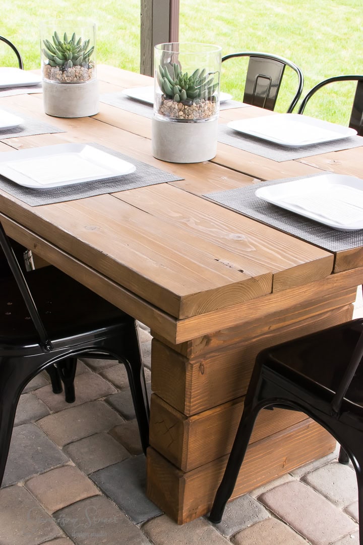 Wooden outdoor dining table set with white square plates, black metal chairs, and two glass centerpieces containing succulents and pebbles. The table is elegantly placed on a brick patio in the backyard, offering a serene grassy background.