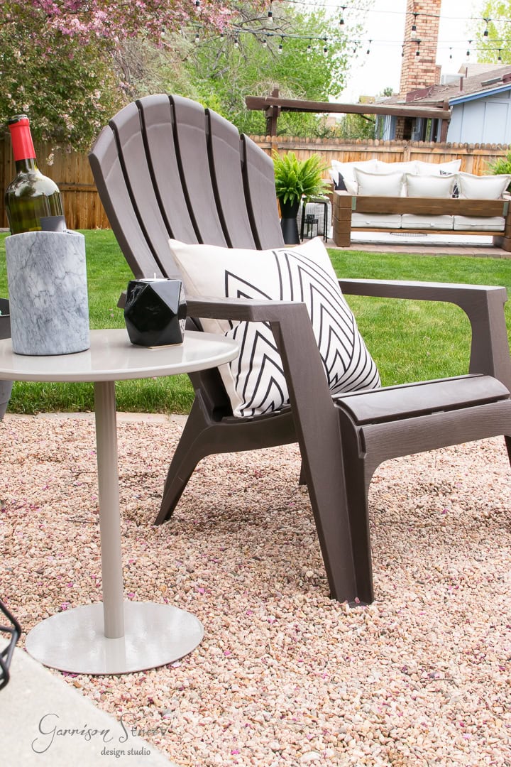 A brown Adirondack chair with a black and white patterned pillow sits on the gravel patio. On the small round side table, a wine bottle, two glasses, a black geometric object, and a marble container add charm to the backyard scene, where a lawn and sofa are visible in the background.