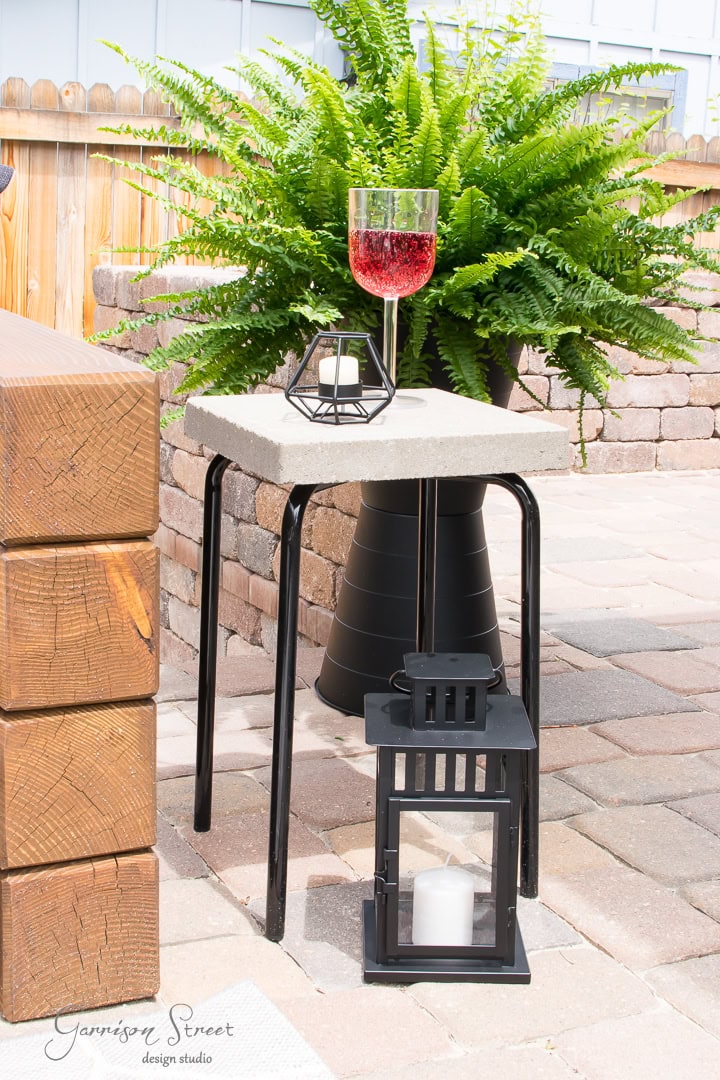 A stylish outdoor setup features a modern Ikea table adorned with a red drink glass and geometric candle holder. A black lantern sits below, complemented by a lush green fern in the background. Brick pathways and wooden elements create a cozy atmosphere.