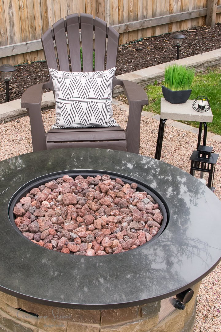 The patio features a gray Adirondack chair with a geometric-patterned cushion, set in an inviting outdoor seating area. A round fire pit filled with red lava rocks takes center stage, while a small side table holds a decorative plant and lantern. Gravel paths and a wooden fence complete this backyard oasis.