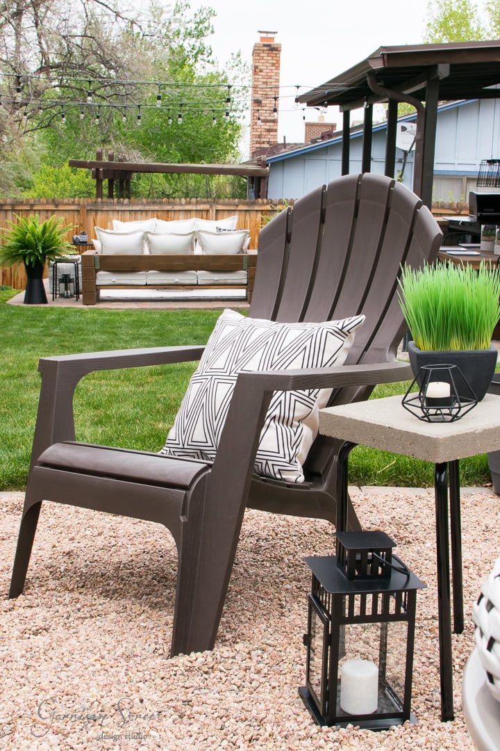 A cozy backyard setup features a brown Adirondack chair with geometric pillows, complemented by a small black lantern and planter on a side table. In the background, there's inviting patio furniture surrounded by green grass and leafy trees under a cloudy sky.