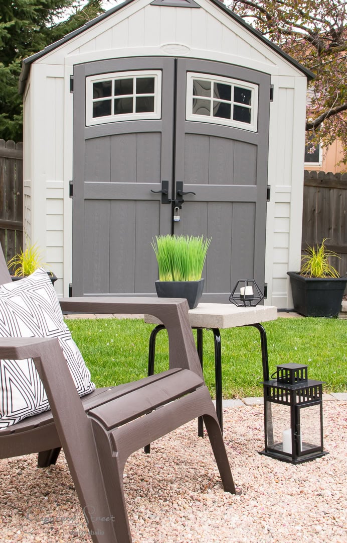 An inviting backyard scene showcases a gray and white garden shed with two small windows. In the foreground, a cozy brown patio chair, a table with a plant, and two decorative lanterns rest on gravel, surrounded by lush greenery and enclosed by a wooden fence.