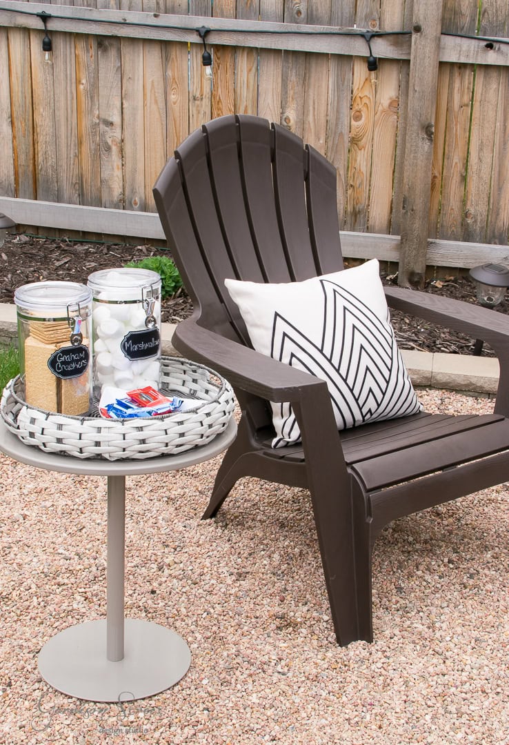 A brown outdoor chair with a black and white patterned pillow sits on gravel in the cozy backyard. Next to it, a small round table holds jars of graham crackers and marshmallows, along with a tray of chocolate bars for making s'mores. A wooden fence completes this serene patio setting.