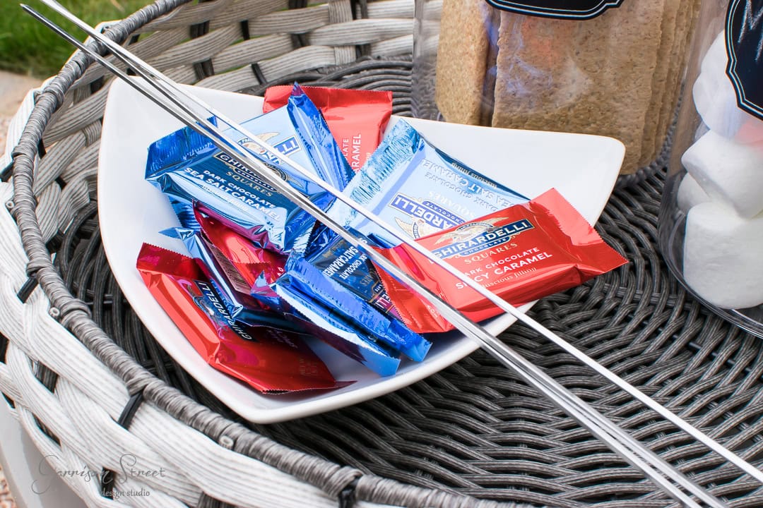 A white bowl filled with assorted Ghirardelli chocolate squares, wrapped in red and blue foil, sits on a wicker surface on the patio next to two metal skewers. Nearby, jars of marshmallows and brown sugar are visible, adding a sweet touch to the backyard setup.