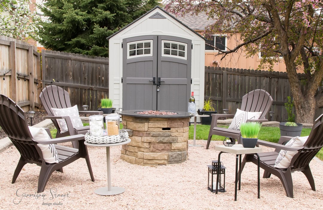 A cozy patio scene with four Adirondack chairs around a round stone fire pit. Two small tables hold plants and lanterns, adding charm to the backyard oasis. A plastic storage shed is in the background, surrounded by a wooden fence and trees.