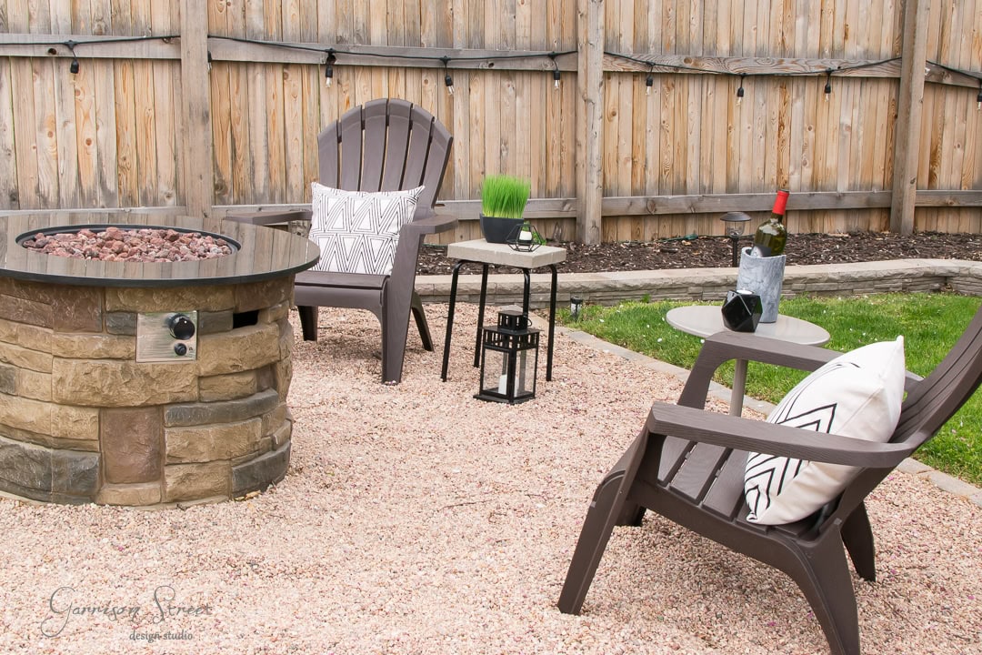 A cozy backyard patio features two brown chairs around a stone fire pit. A small table holds a potted grass plant and two wine glasses, while a patterned pillow decorates one chair. A wooden fence encloses the gravel area, crafting an inviting atmosphere.