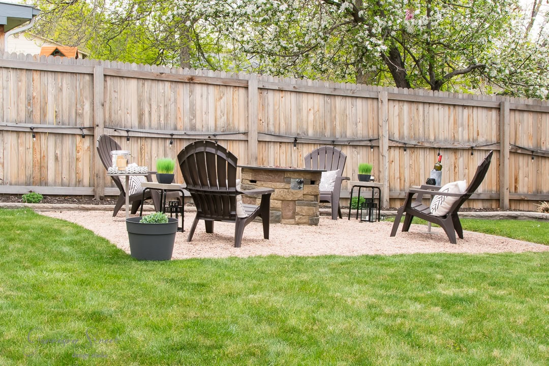 A cozy patio scene features four Adirondack chairs around a stone fire pit on a gravel square. Two side tables hold potted plants and decorative items. A large tree with blooming flowers and a wooden fence provide a charming backdrop, with grass surrounding the backyard area.