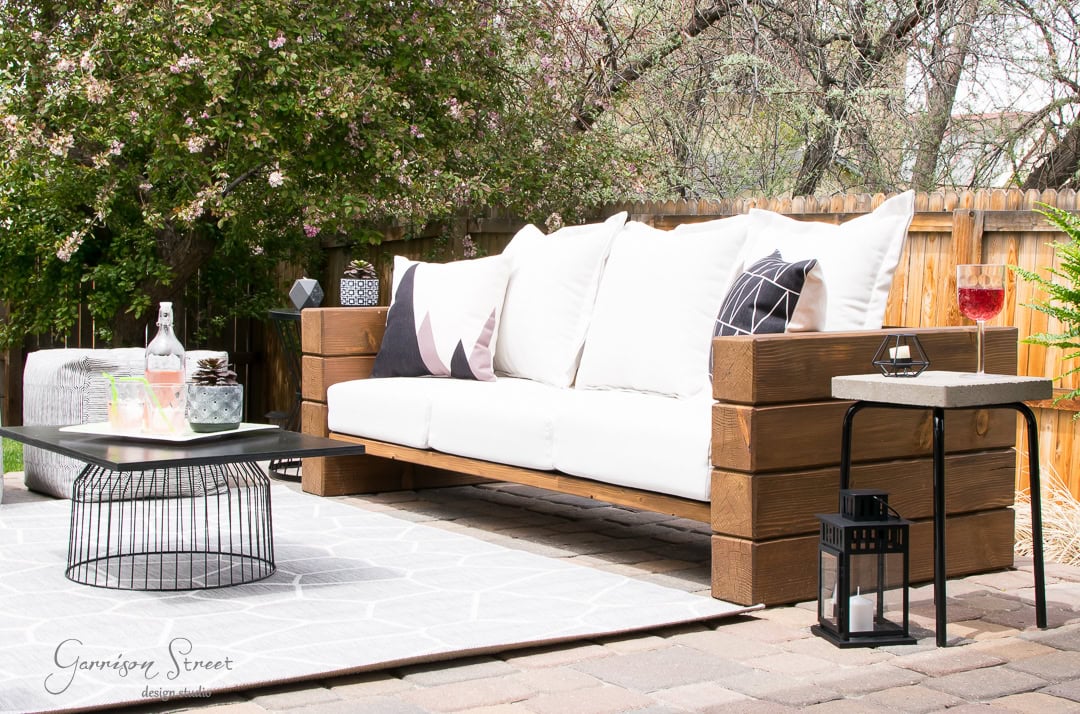 A serene backyard patio hosts a wooden sofa adorned with white cushions and patterned pillows. A geometric wire-frame coffee table holds decor items, while a lantern sits on a side table beside the sofa. Wine glasses grace the tables, surrounded by lush greenery.