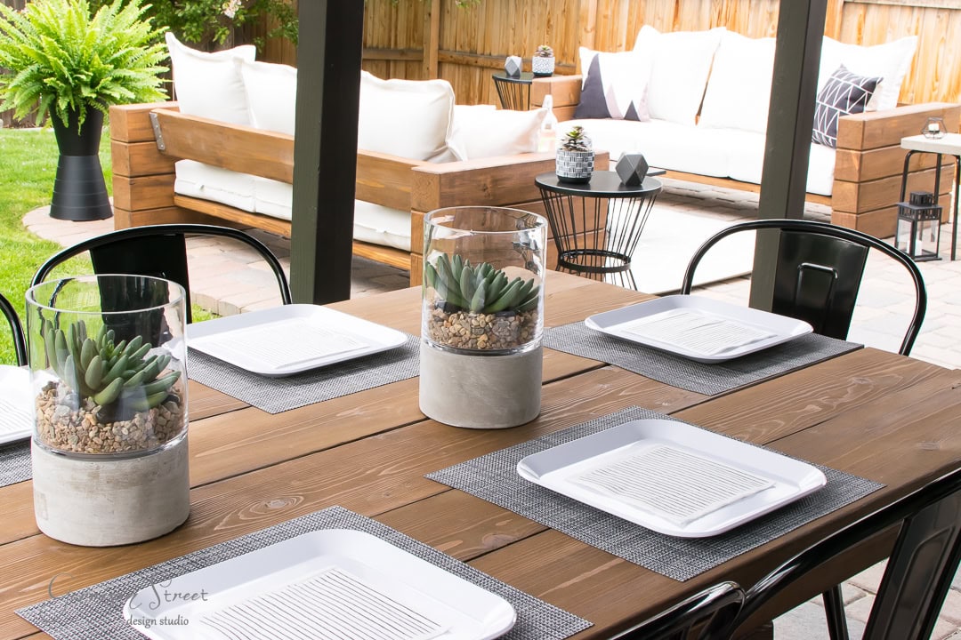 Outdoor dining area in the backyard with a wooden table set for four, featuring white plates on gray placemats. Decorative glass containers with succulents adorn the table. In the background, a cozy patio with a white sofa and lush plants is visible.