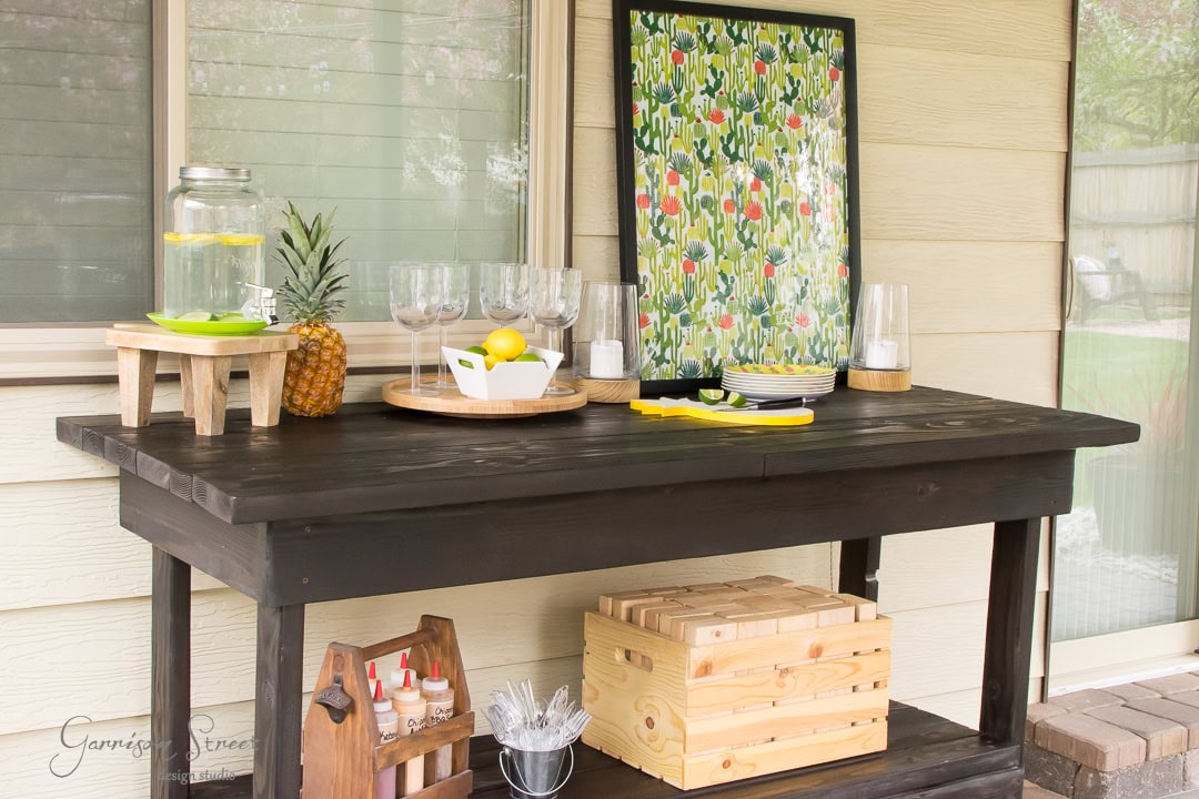 A black outdoor table on the patio holds a lemonade dispenser with glasses, a pineapple, lemons on a tray, and a colorful floral painting. Below, a crate with jars and utensils is visible, completing this charming backyard setup against the backdrop of house siding.