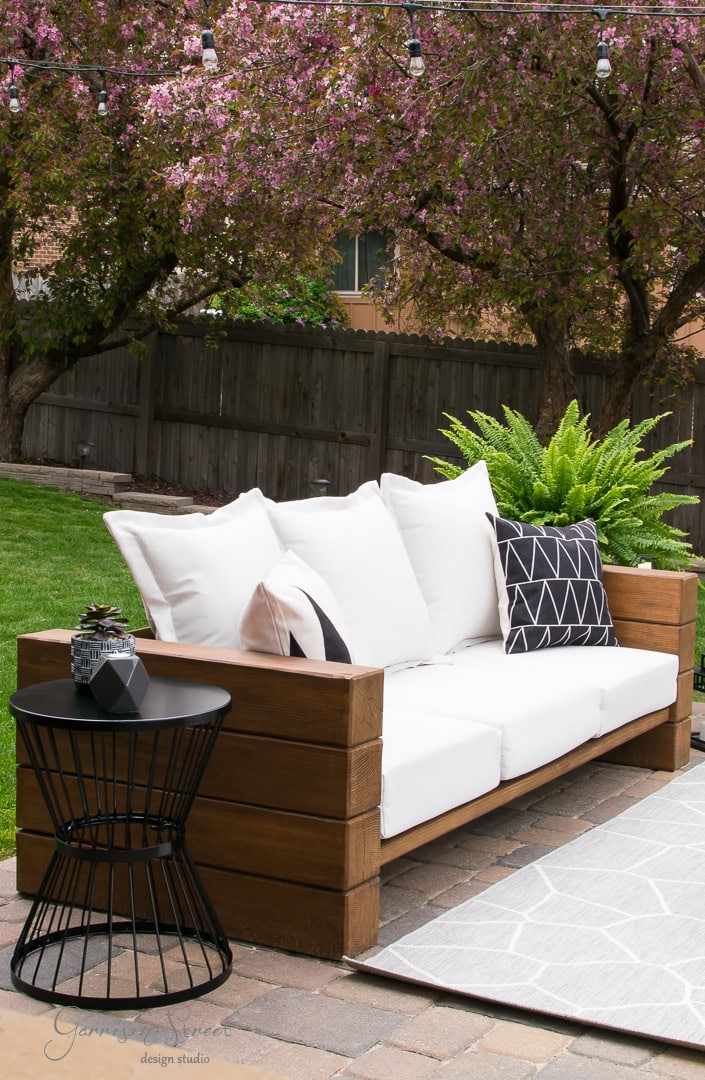 A cozy backyard patio scene showcases a wooden sofa adorned with white cushions and decorative black and white pillows. Nearby, a black side table holds a lantern, while flowering trees and string lights in the background add a charming touch.