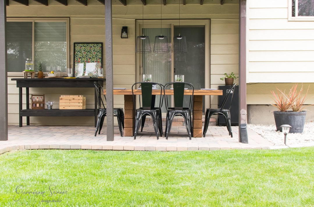 The backyard offers a delightful outdoor dining area featuring a wooden table, black metal chairs, and a sleek serving cart. The covered patio is surrounded by a small grassy yard, with hanging lights and plants adding a cozy touch to the ambiance.
