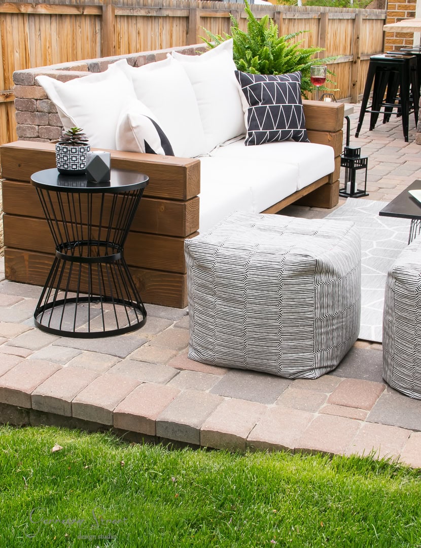 A cozy backyard retreat features a brick patio with a wooden sofa adorned with white and black geometric cushions. A round black metal side table and two gray square poufs provide additional seating, while a wooden fence and lush green grass complete this inviting outdoor space.