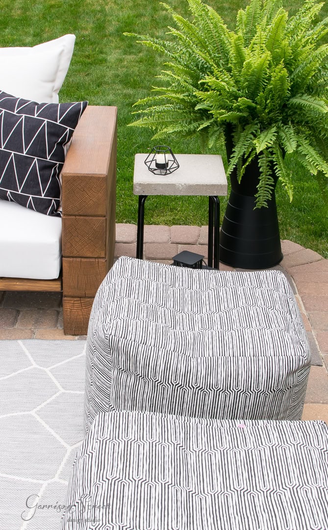An outdoor patio setting features Ikea geometric-patterned ottomans, a wooden armchair with a black-and-white cushion, and a small table showcasing a clever hack – a geometric candle holder. A lush green fern and a light gray rug cover the stone patio floor.