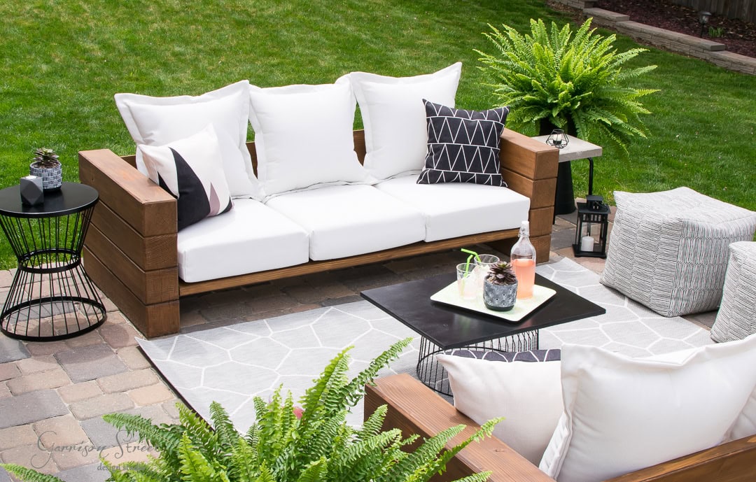 Home Outdoor patio and backyard retreat featuring a wooden sofa with white cushions, geometric black and white pillows, a black coffee table with drinks, potted ferns, a small side table, and a gray and white patterned rug on the brick surface. A lush green lawn lies beautifully in the background.