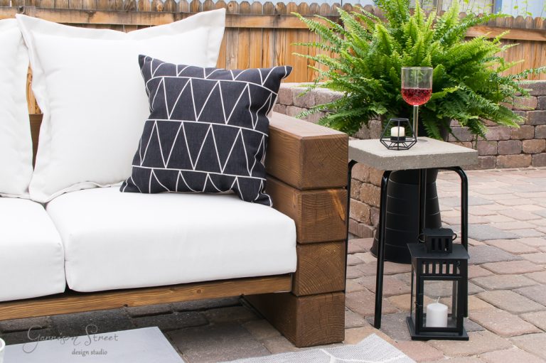 Outdoor patio with an Ikea wooden sofa featuring white and patterned black cushions. A small concrete table holds a wine glass and decorative lantern. A lush fern sits behind the table, all set on a stone patio with a brick fence.