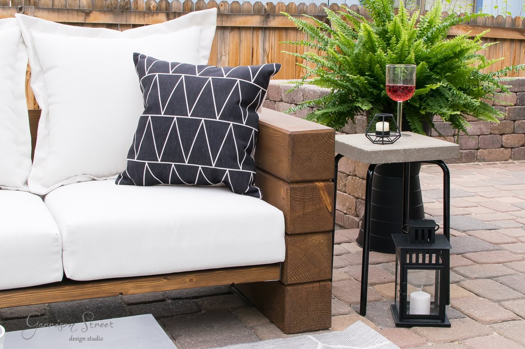 Outdoor patio with an Ikea wooden sofa featuring white and patterned black cushions. A small concrete table holds a wine glass and decorative lantern. A lush fern sits behind the table, all set on a stone patio with a brick fence.