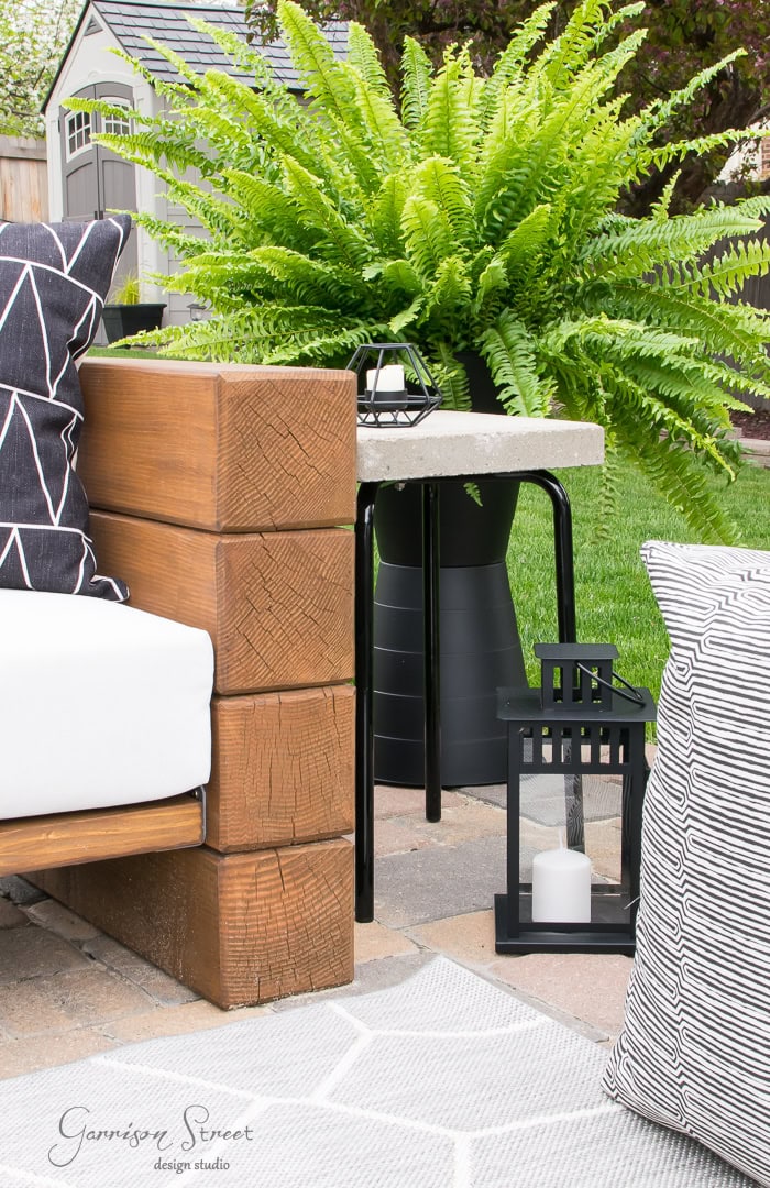 In the backyard, the outdoor patio scene comes to life with a wooden bench adorned with black and white patterned cushions. A large green fern nestles in a black planter beside a black lantern casting candlelight on the stone floor. The backdrop features a rustic wooden fence and shed.