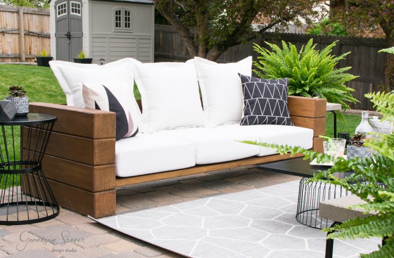 A stylish outdoor patio in the backyard features a wooden sofa with white cushions and geometric-patterned pillows. A gray rug lies beneath, and a lush green fern sits beside the sofa. In the background, there's a small garden shed and a fenced yard.