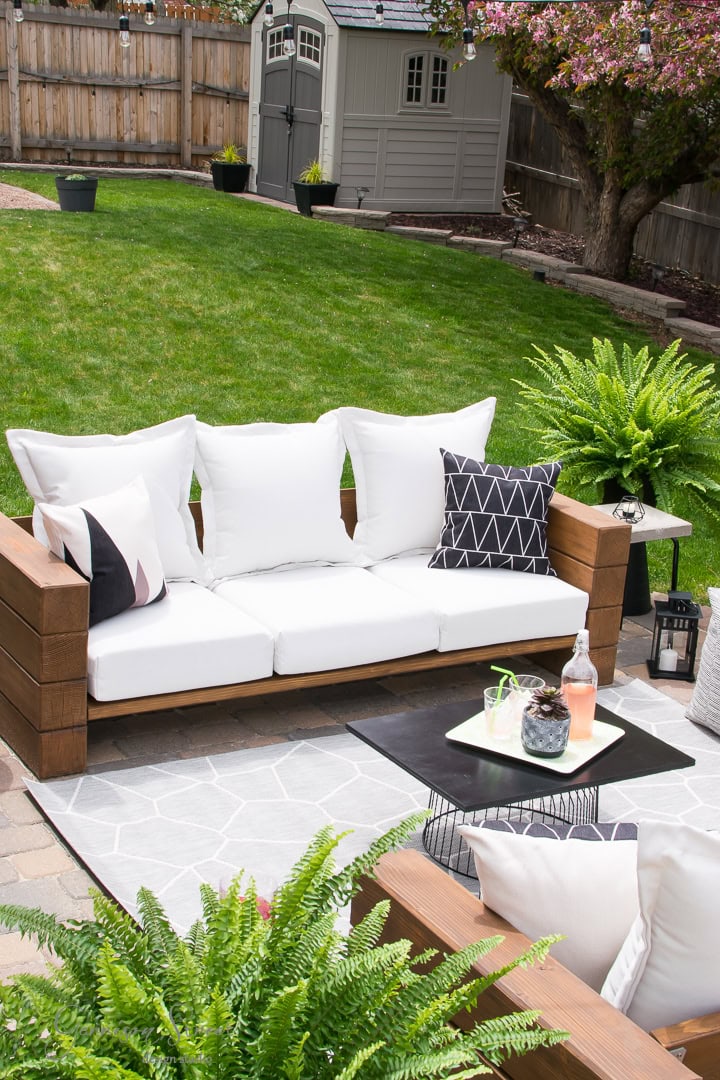 The backyard patio boasts a wooden couch adorned with white cushions and geometric pillows. A black coffee table holds drinks and snacks, while a lush lawn, potted ferns, and a blooming tree cultivate a serene garden atmosphere. In the background, a small shed quietly stands amid nature’s embrace.