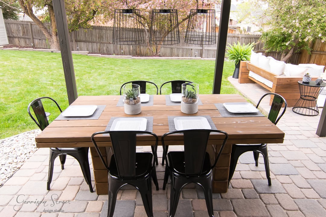 A wooden outdoor dining table set for six with black metal chairs sits on a stone patio in the backyard. The table is adorned with potted plants and square plates. In the background, there's a grassy lawn, a fence, and a patio sofa.