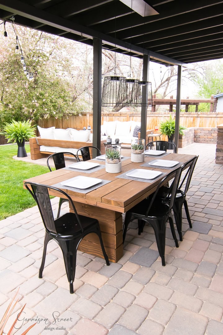 A charming backyard patio showcases a wooden dining table set for six, with black metal chairs and white plates. A decorative centerpiece plant graces the table. String lights and hanging lamps adorn the covered pergola, while a bench and lush greenery complete the inviting space.