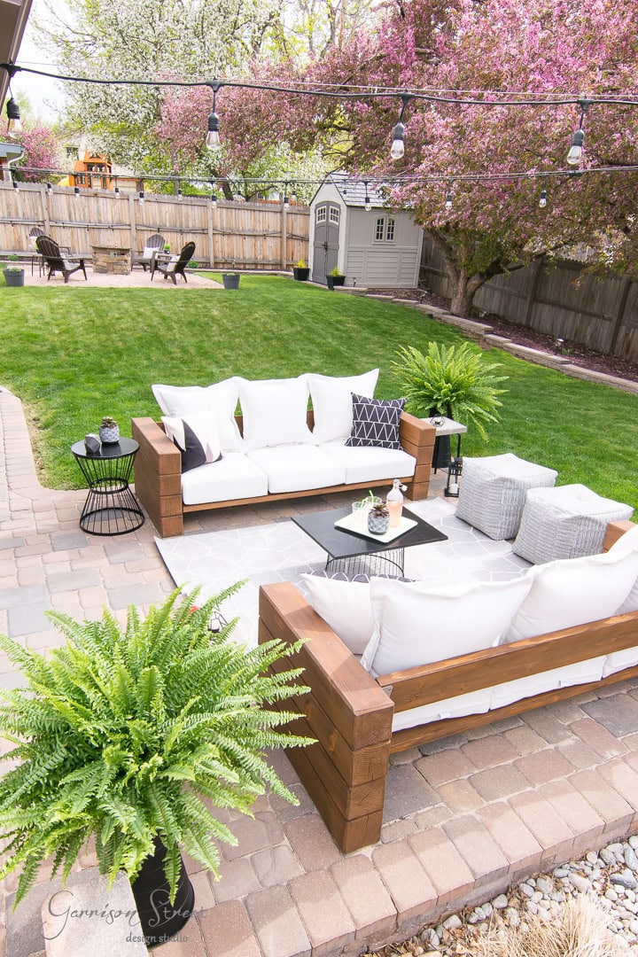 A backyard patio showcases a DIY outdoor sofa with white cushions next to a sleek black coffee table. Potted ferns adorn the area, while in the background, a grassy lawn, a white shed, trees with pink blossoms, and string lights enhance the cozy atmosphere.
