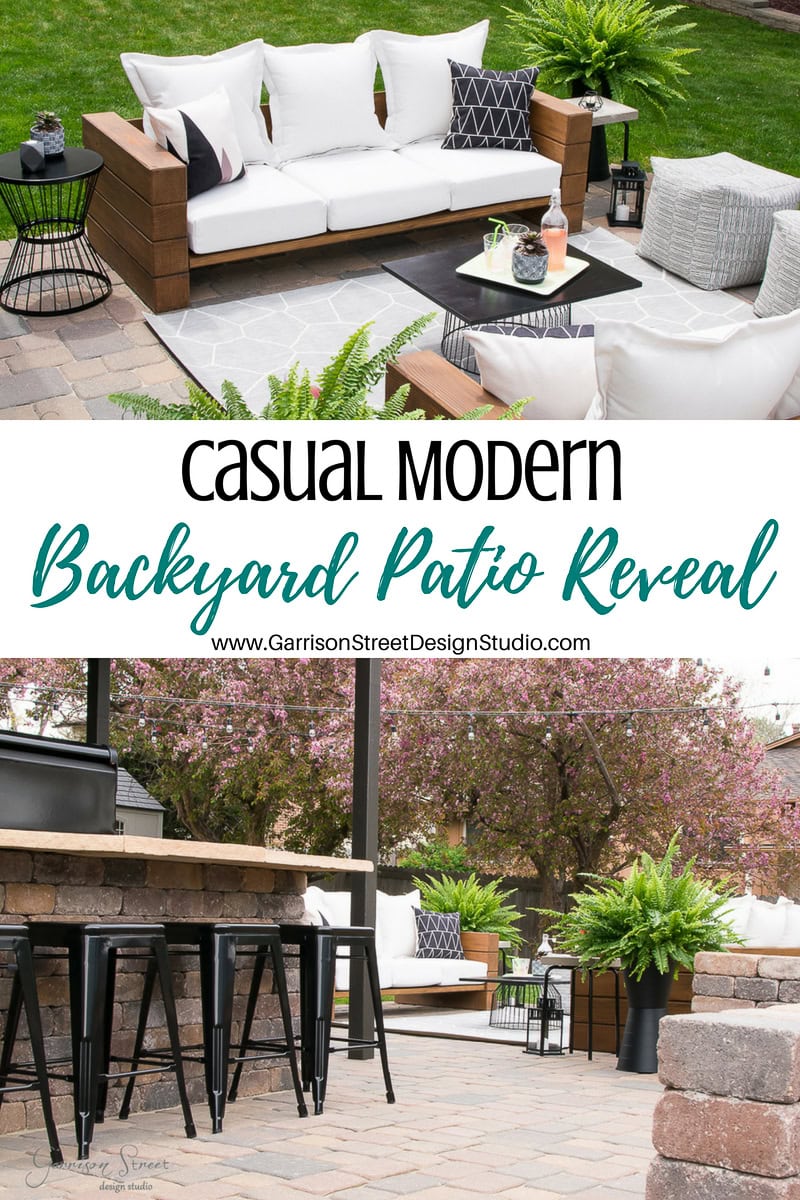 A modern backyard patio showcases a white sofa with a wooden frame, black and white cushions, and a sleek black coffee table. This serene setting includes a bar with stools, a brick fire pit, and is enveloped by lush greenery and pink flowering trees.