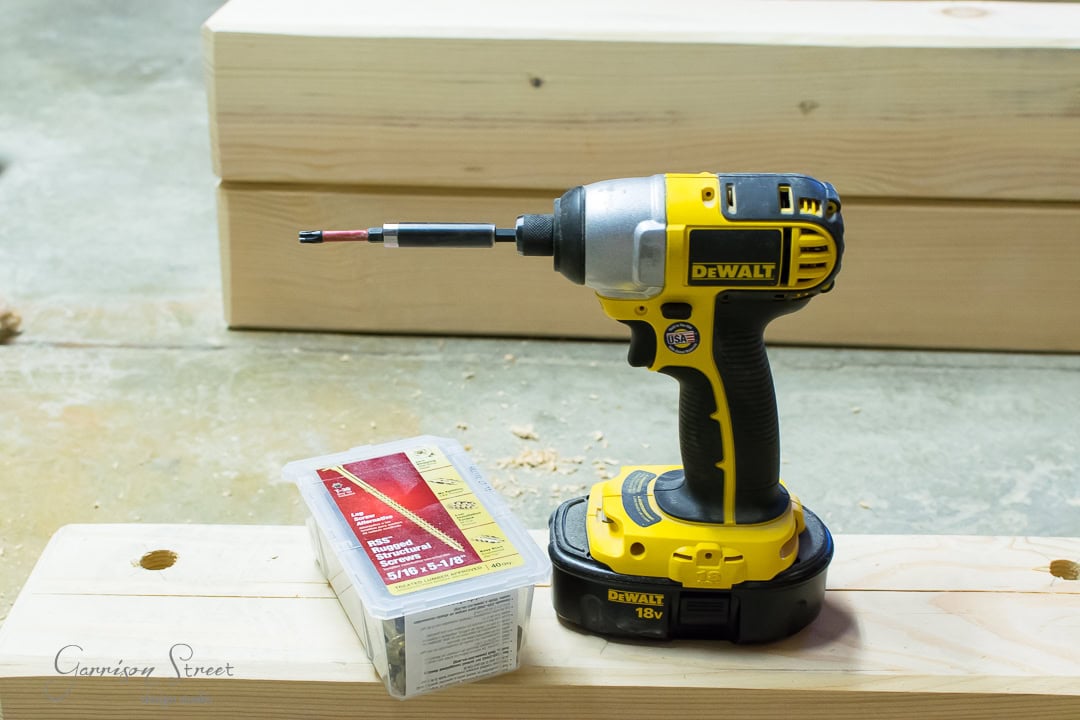 A yellow DeWalt 18V cordless impact driver sits on a wooden board, ready to conquer your next project. Nearby, a box of screws waits its turn, set against a backdrop of stacked wooden beams.