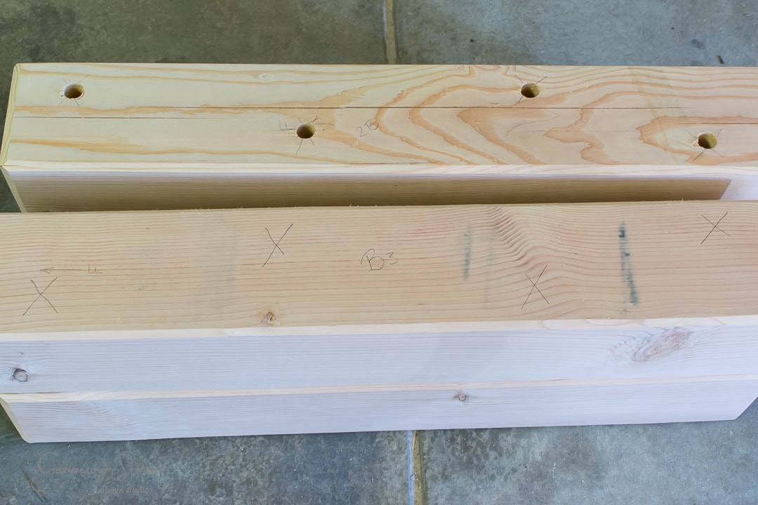 Two wood beams lie on a concrete floor, marked with pencil lines and X's. The top beam features four circular holes, showcasing the precision needed for this project, while both beams display their natural wood grain patterns beautifully.