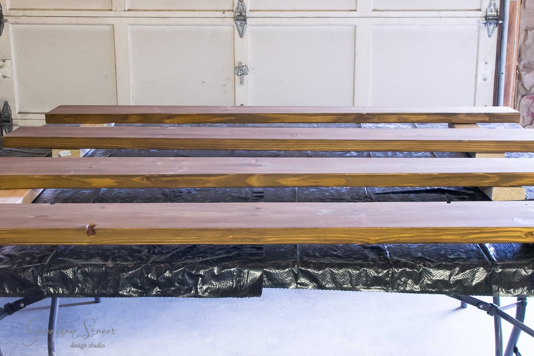 Wooden planks laid out on a metal table in a garage setting, ready for staining or finishing. The background shows a white garage door with metal hinges.