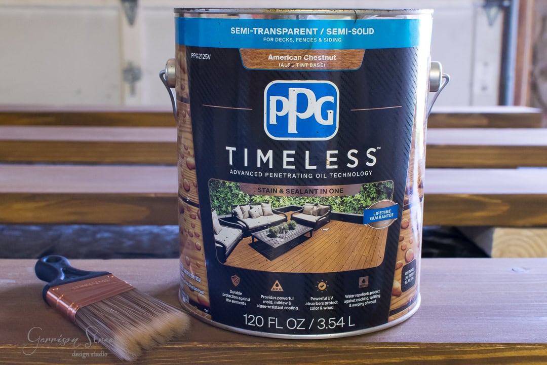 A paint can labeled "PPG Timeless" in rich American Chestnut, boasting a semi-transparent finish, rests on wooden planks beside an outdoor sofa. Nearby, a paintbrush lies ready. This can promises stain and sealant in one, backed by advanced technology for lasting beauty.