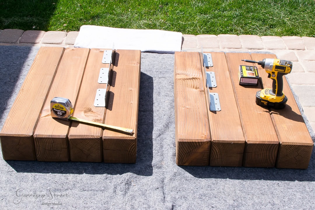 Two wooden platforms with metal brackets are set on a gray surface outside. A tape measure rests on the left platform, while a yellow drill with screws sits ready on the right. Brick paving and patches of grass complete the scene.