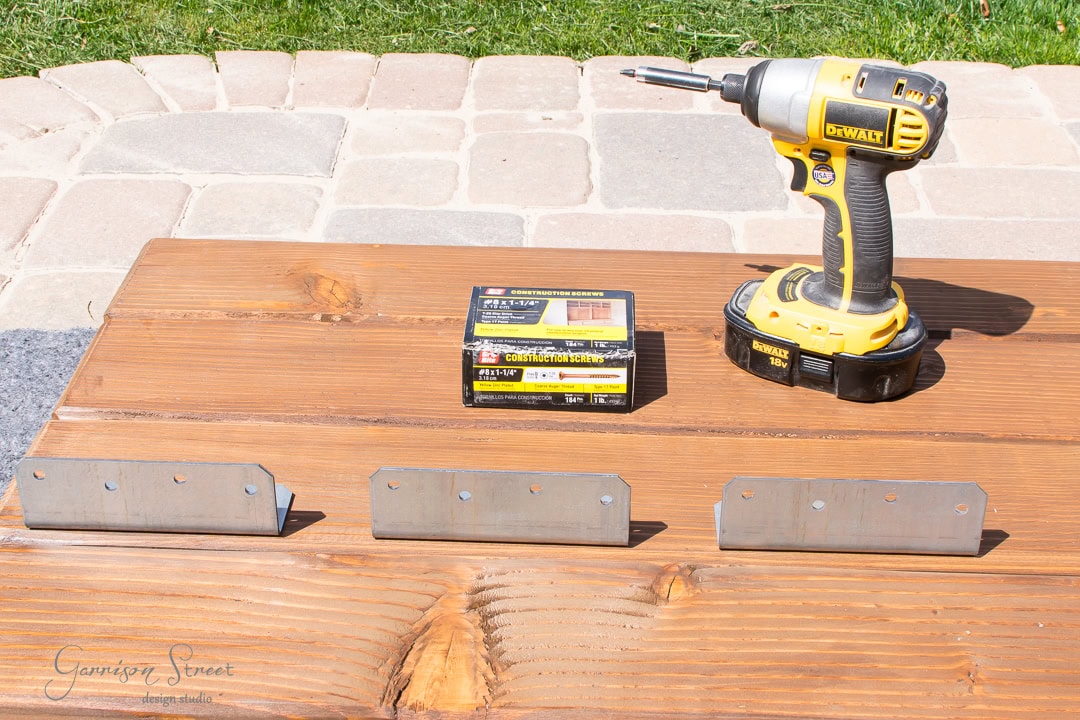 A wooden table outdoors displays a power drill, a box of wood screws, and three metal brackets. The backdrop features a stone patio and lush grass, setting the perfect scene for crafting your garden furniture.