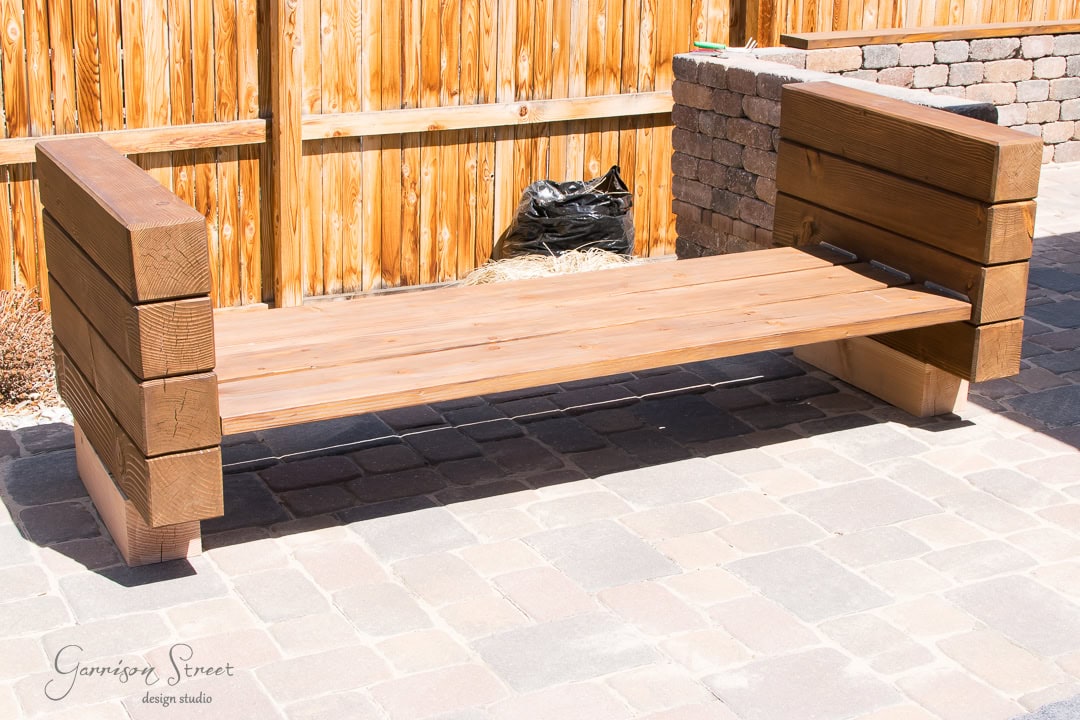 A wooden outdoor bench sits gracefully on a stone-paved patio surrounded by a wooden fence. The bench boasts a simple, sturdy design with thick timber planks, while a black plastic bag lingers subtly in the background.