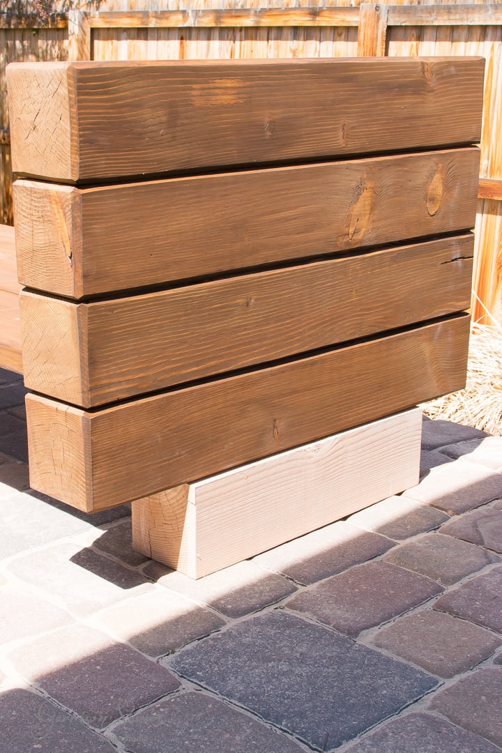 A wooden bench with a simple design made of thick horizontal planks is placed on a stone-paved surface. The bench has a natural finish, and the backdrop features a wooden fence.