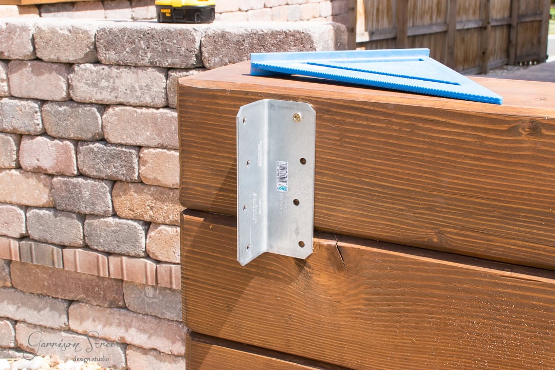 Close-up of a metal bracket attached to a wooden beam with screws. A blue plastic square tool rests on the beam. In the background, there's a wall made of beige and red bricks.