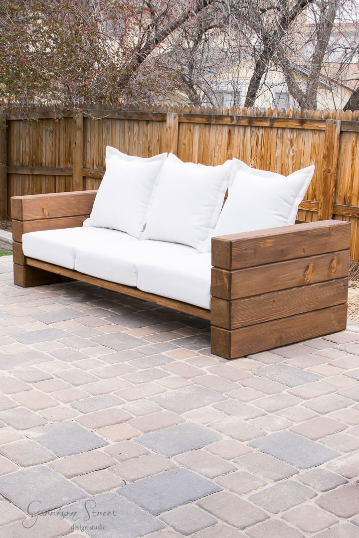 A DIY outdoor sofa with thick white cushions and pillows sits elegantly on a stone patio. Its wide, sturdy armrests invite relaxation. In the background, a wooden fence and leafless trees create a serene setting.